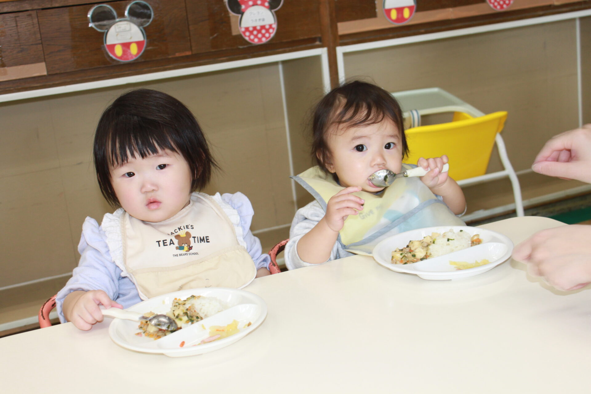 スタイを付けた幼児二人がスプーンで食事をしている写真